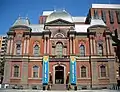 A 3-story, brick and red stone, second empire style building with a slate roof. The building is symmetrical with the entrance center and a blue and yellow banner on either side.
