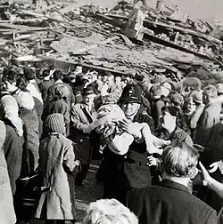 A policeman carries a girl. They are surrounded by a crowd, and there is a large pile of rubble in the background.
