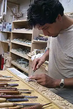 Man carving a strip of wood with carving tools lined up in front of his workstation