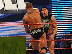 Two dark haired men standing in a wrestling ring with red ropes, one with his back to the camera. The man with his back to the camera is wearing short black wrestling tights with the name 'Orton' written on them in white lettering, and has a tribal design tattoo on his back. He is throwing a punch at the other man, who is facing the camera with a look of surprise, and is wearing short purple wrestling tights and a black top with a white design on it.