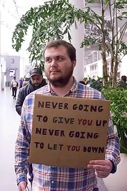 A person holds a cardboard sign with the words, "Never going to give you up, Never going to let you down".
