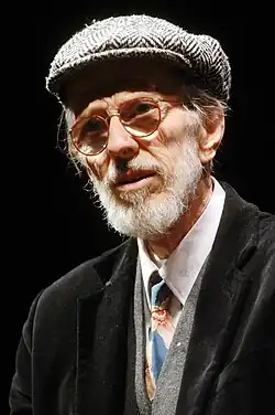 An elderly man with a white beard, round glasses, and flat cap.
