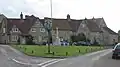 Street scene. Triangular area of grass with village sign on wooden post and stone cross behind. Stone houses with tiled roofs n the background.