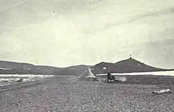 A conical tent stands on a desolate stretch of beach, with a range of low hills in the background.