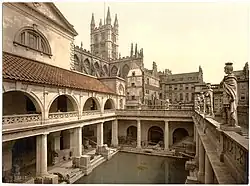 A late-nineteenth-century Photochrom of the Great Bath at the Roman Baths. Pillars tower over the water, and the spires of Bath Abbey – restored in the early sixteenth century – are visible in the background.