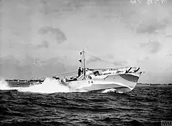 Ship speeding at sea with a white bow wave; land can be seen in the background