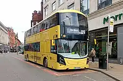Three yellow buses lines up such that the front on is just right-of centre and they are at an angle with the third one going off screen. They are at a bus stop with tall buildings all around and someone waiting outside the first bus; the photo is taken from across the road.