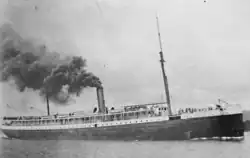 A grainy black and white photo of a steamship on the water. The bow faces to right and the ship occupies the whole frame. Several groups of people are visible fore and aft, on the first and second deck. The ship has two masts and a smokestack which is bellowing.