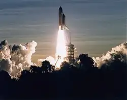 A space shuttle launches into a dawn sky. Clouds in the sky, in the launch plume and from the flame trench, are visible, as is the scaffolding-like launchpad and some vegetation silhouetted in the foreground.