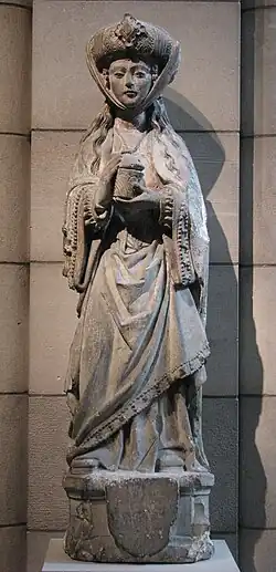 St. Mary Magdalene, wearing a jeweled hat and wearing a crucifix and ornate robes, holds an open ointment jar. A sculpture carved of limestone.