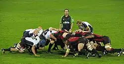 Two opposing formations of eight men, in white and black to the left, red and black to the right, push against each other in a crouched position; behind them stands another player and the referee