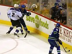 Two ice hockey teammates in the process of passing to one another, while an opposing player checks one of them into the boards. The teammates are dressed in a blue jersey, while the opposing player is dressed in white.