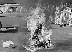 Vietnamese Mahayana Buddhist monk Thich Quang Duc during his self-immolation at a Saigon road intersection. Quang Duc is sat down in the traditional Buddhist meditative lotus position, maintaining an outward composure while flames consume his robes and flesh, and black oily smoke emanates from his burning body. Also appearing in the frame: dozens of monks and nuns watching the scene, an Austin Westminster sedan a few meters behind Quang Duc, and an empty five-gallon petrol container next to him.