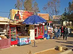 Two small shops offering Coca-Cola products and other soft drinks in glass-fronted refrigerated containers. In the middle is an umbrella with the Pepsi logo. Above them are signs referencing Indiana Jones and depicting him. A small group of people is walking past and in front of them on the right
