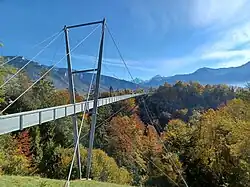 Suspension footbridge spanning a forested gorge in a mountainous landscape, with steel support towers, cables, and autumn-colored trees under a clear blue sky.