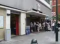 A modern looking building with marble coloured tiled wall at ground level with grey cladding above. A small concrete canopy is over the larger of two entrances with blue signs reading "SLOANE SQUARE STATION", people are entering and exiting through the larger entrance.