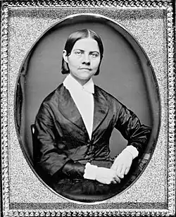 Framed monochrome photograph portrait of a woman sitting, shown from the waist up, left elbow resting on furniture, hands together in lap, the woman wearing a black silk jacket which narrows to conform to the waist, bearing curved lapels, over a plain white blouse with a collar closed at the throat. The woman has dark, straight hair parted in the middle and cut short at the top of the collar. Her head is tilted slightly to her left, face forward, and she is looking directly at the observer.