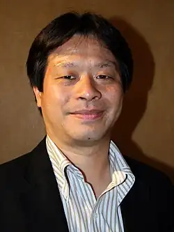 Portrait of a Japanese man smiling at the camera wearing a black suit jacket and a gold striped dress shirt.
