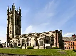 A big church as photographed from slightly lower down the hill, such that the long side of the cross shape is runs the length of the photo, with the spire on the left-hand side. The church resembles an L shape because the spire is right at one end and the three other parts of the cross are very small.