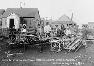 Ten men stand behind an array of empty dog sleds, some on racks. Most are Inuit and wear traditional or western working clothes: one is Caucasian and is dressed as a clergyman.