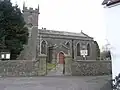 Gray bstone building with square tower to the left hand end, partially obscured by tree. In the foreground is a stone wall separating the church from the road.