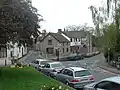 Street scene showing road junction and grey stone buildings with parked cars in front of them. To the left is a grassy area with a tree.