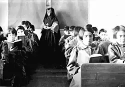 Students in the classroom, with a teacher in nun's garb at the back of the room.