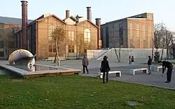 A brick factory stands in front of a park, with open green space, a reflecting pool, and benches