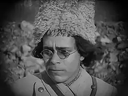 Still from a movie showing an actor dressed as Nestor Makhno, wearing glasses, a cossack military uniform and a ushanka