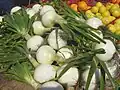 A pile of onions, with green stems protruding from white bulbs. Organes, lemons and limes are visible in the background.
