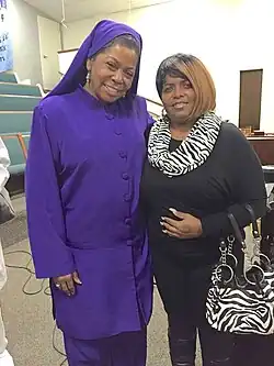 Two middle-aged black woman stand looking at the viewer, evidently posing for the photograph. The woman on the left is wearing a purple outfit with long sleeves and a matching headpiece.