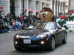 A black open-top sports car with lights on is being driven down an asphalt road. A large furry toy costume, slightly larger than a human is standing in the back seat. It has cream coloured mouth and chest, and dark brown arms and forehead, large whiskers, a grin, large white eyes and two canines. Behind him are some men walking in green costumes. On the left is a crowd watching the parade from the footpath, in front of tall buildings with stone arches.