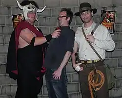 A photograph of three individuals, two in fancy dress. The individual on the left is wearing a cow skull headpiece and pretending to rip out the heart of the person next to him.