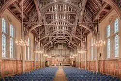 The interior of a grand hall as photographed from the back-centre. The hall is full of chairs and has a grand and intricate beamed roof with chandeliers hanging from the beams and gothic-shaped windows either side