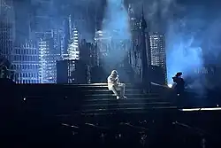 An extreme wide shot of the Weeknd performing with a durag in white-colored fashion as he sits down and performs on the staircase of the concert's setup.