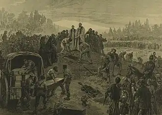 A vicar stands at an open grave, which is being filled with a coffin. Several others are being unloaded from a covered waggon. Crowds of people are shown paying their respects.