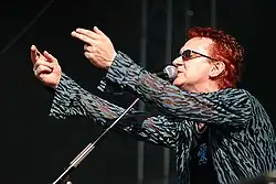 A color photograph of a man, facing him from his left side, on stage at a concert, singing into a microphone and pointing his fingers at the crowd.