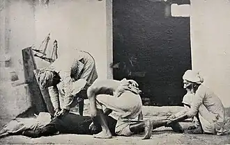 An individual hunched over someone lying face down, holding a cloth around their neck, while two others hold their torso and feet in place