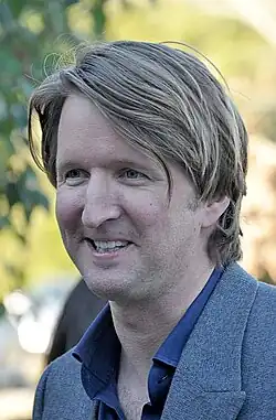 A man in a grey suit with a blue shirt looks down to his right while smiling.