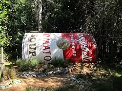 A large Campbell's Tomato Soup can resting on its side in front of trees with shadows of other trees falling on it.