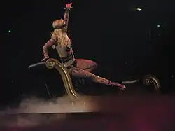 Image of a blond woman. Her right hip is resting on a black divan, with her right leg extended to the end of the divan. Her left hand is extended in the air, while her right hand is grabbing the top of the divan. She is wearing a flesh-colored body suit with tattoos and nipple tassles. She is sporting a black blindfold and singing into a wireless microphone. The divan is surrounded by smoke.