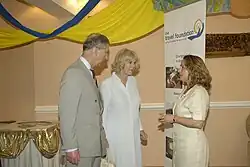 Charles wearing a suit converses with a woman in a dress. Queen Camilla stands next to Charles in the background standing opposite a "Travel Foundation" vertical banner.