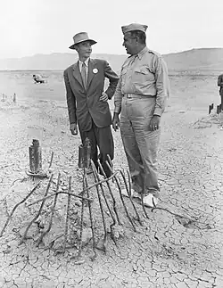 A man smiling in a suit in suit and one in a uniform chat around a pile of twisted metal.