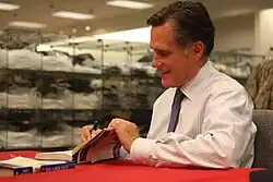 Casual photograph of Mitt Romney indoors seated and signing books