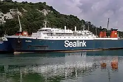 A ferry that is moored viewed from across the water. Its hull is painted blue with "Sealink" in a serif font on top. In the background are chalk cliffs with much vegetation