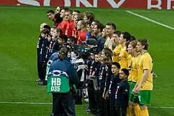 Celtic (foreground, in yellow) and Manchester United players (background, in red) and the match officials (in grey) line up prior to the group stage match between the two sides. Each player has a child mascot wearing a navy blue shirt standing in front of him, and a television camera crew is focusing on the Celtic players.