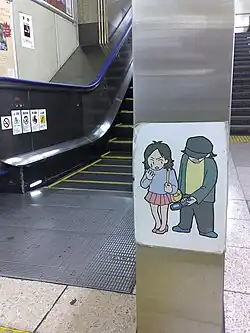 On a metal pillar in front of an escalator, a paper laminated sign shows a graphic of someone in heels and a short skirt sweating and looking backward, upset, at someone whose face is covered by their hat and is taking a picture up the first person's skirt with a flip-phone.