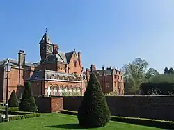 An irregularly shaped red-brick house with slate roofs with an orangery on the ground floor and gables, chimneys and a clock tower above. In the foreground is a formal garden with a lawn, clipped shrubs, a wall and a Victorian-style lamppost.