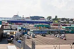 In this photograph, many large containers and other cargo are lined up in the city's ferry port. A ferry can be seen docked in the background.