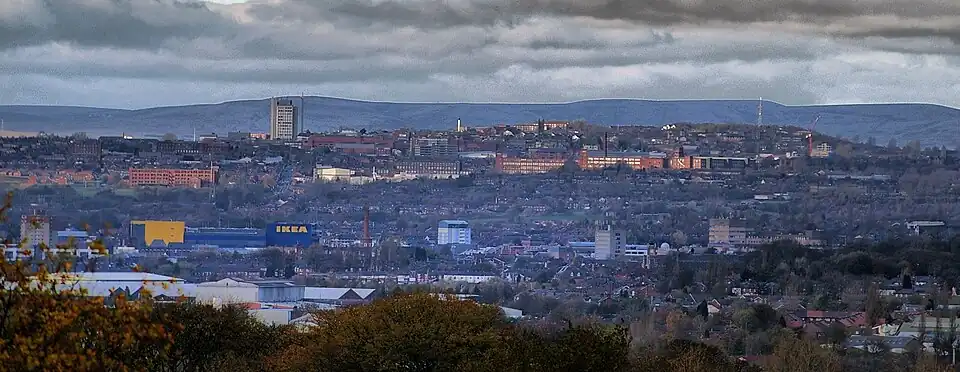 A panoramic shot of a town at the bottom of a hill, and another about halfway up the hill, although they seem fairly contiguous to one another. On the bottom, obvious buildings from left to right are an IKEA, the chimney of the mill, and three brutalist tower blocks. On the top, obvious buildings from left to right are a brutalist tower block, some renovated mill buildings looking over the hillside, and a transmitting tower at the top of the hill.